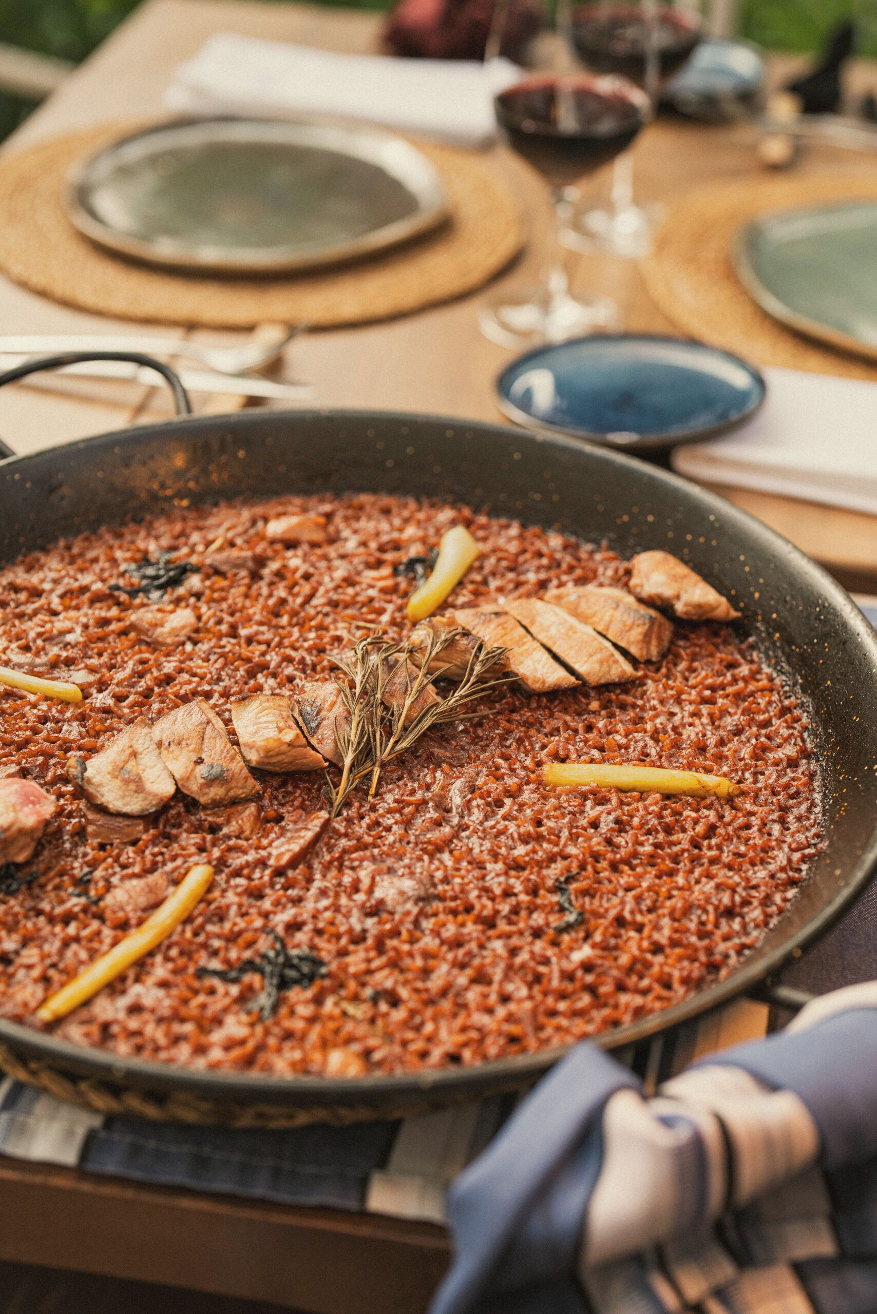 A large pan filled with a dish, possibly rice, garnished with herbs and lemon slices, placed on a table with plates and a glass.