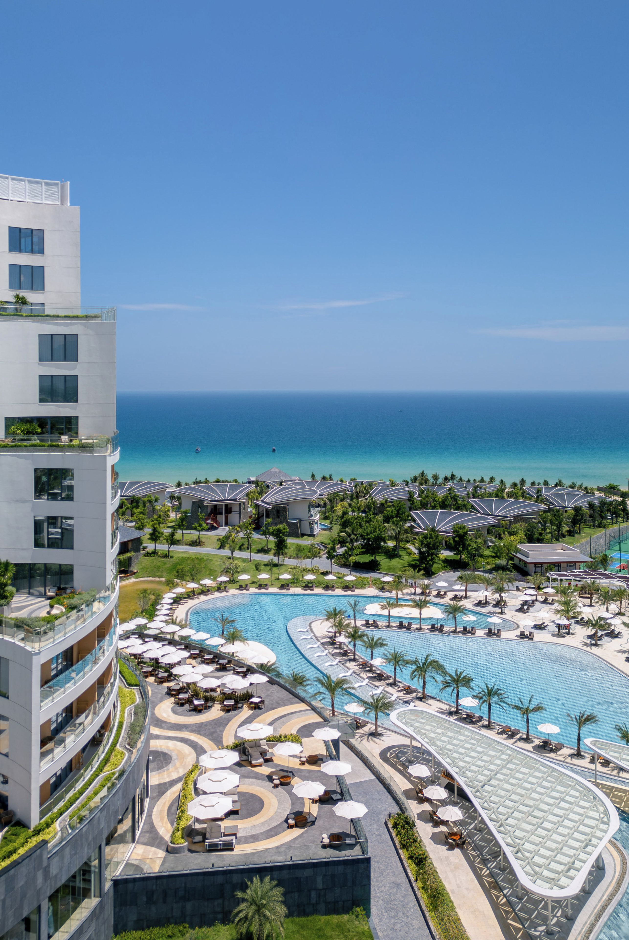 Ein Resort mit großem Pool, mehreren Sonnenschirmen und Blick aufs Meer.