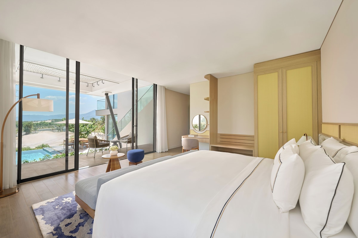 Habitación de hotel moderna con una cama grande, vista a una piscina y zona exterior, y puerta corrediza de vidrio.