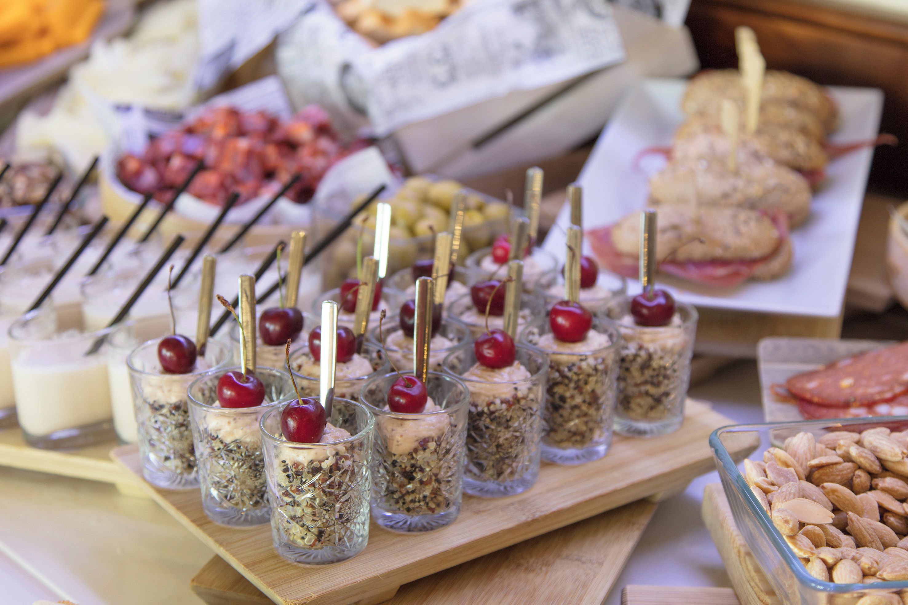 A variety of food items including desserts in glasses with cherries, a tray of sandwiches, and a bowl of almonds.