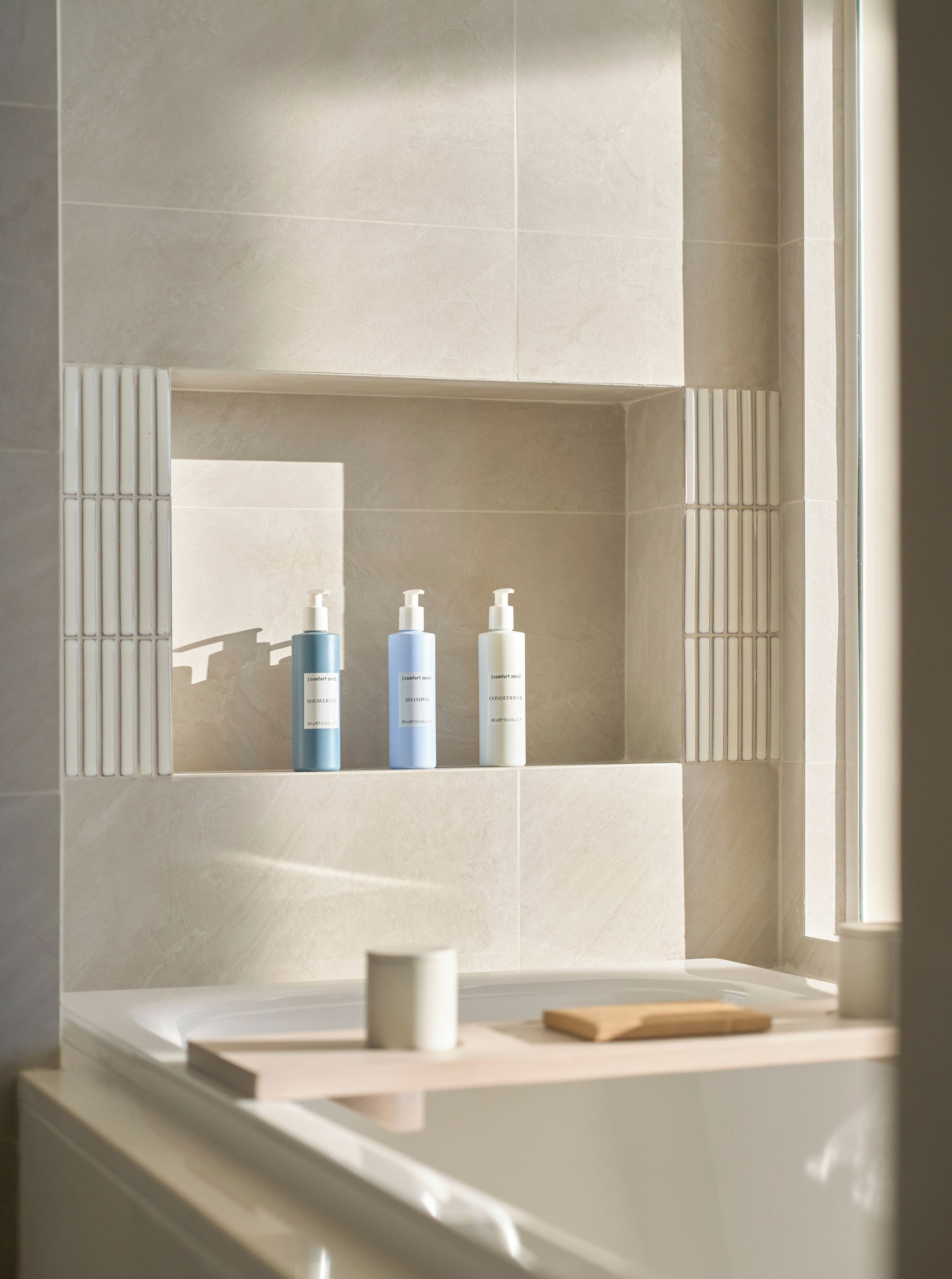 A bathroom with a large mirror, three bottles of lotion, and a wooden bath mat on the counter.