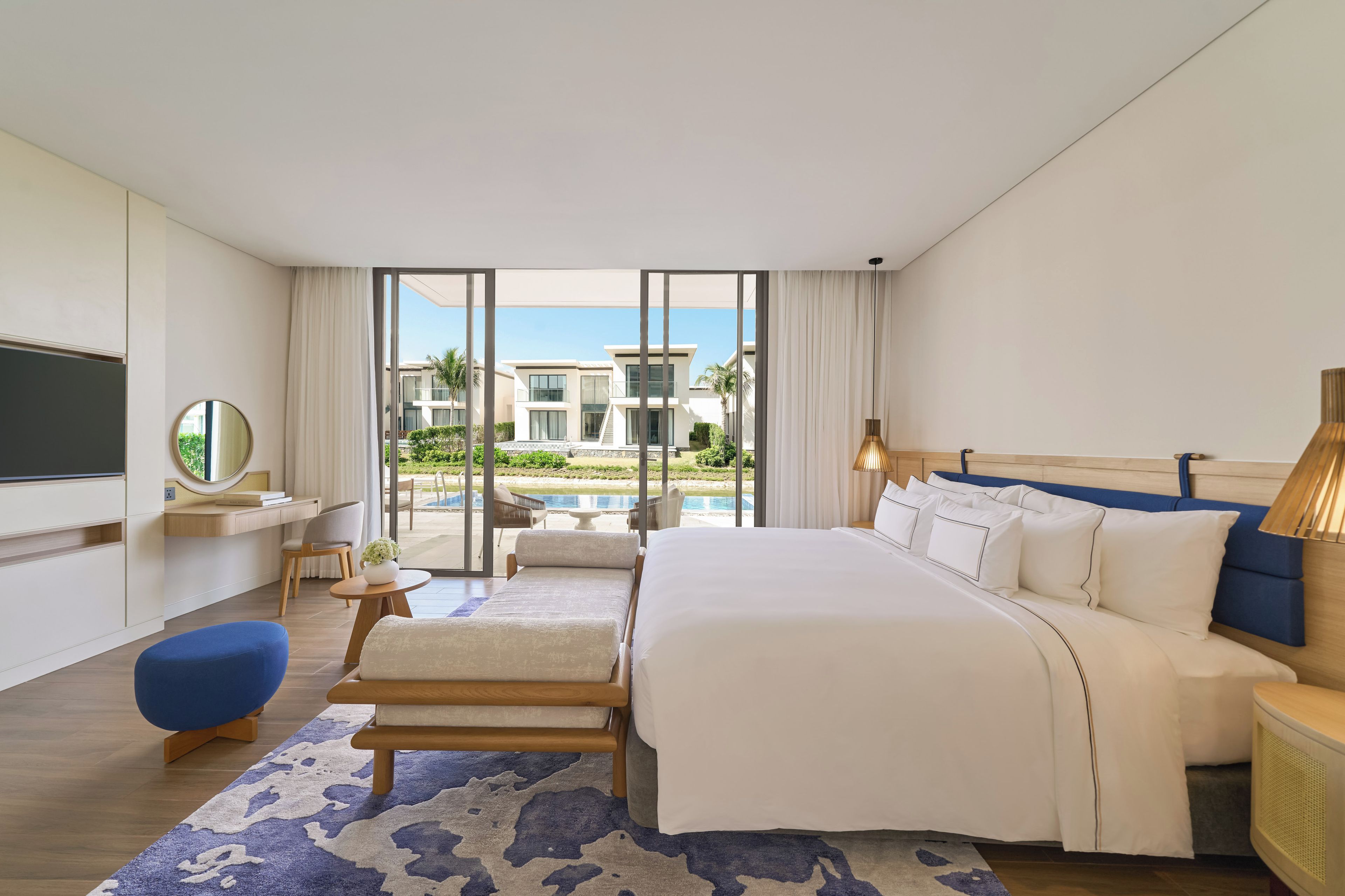 A modern hotel room with a large bed, a TV, a desk, and a view of a pool and buildings.