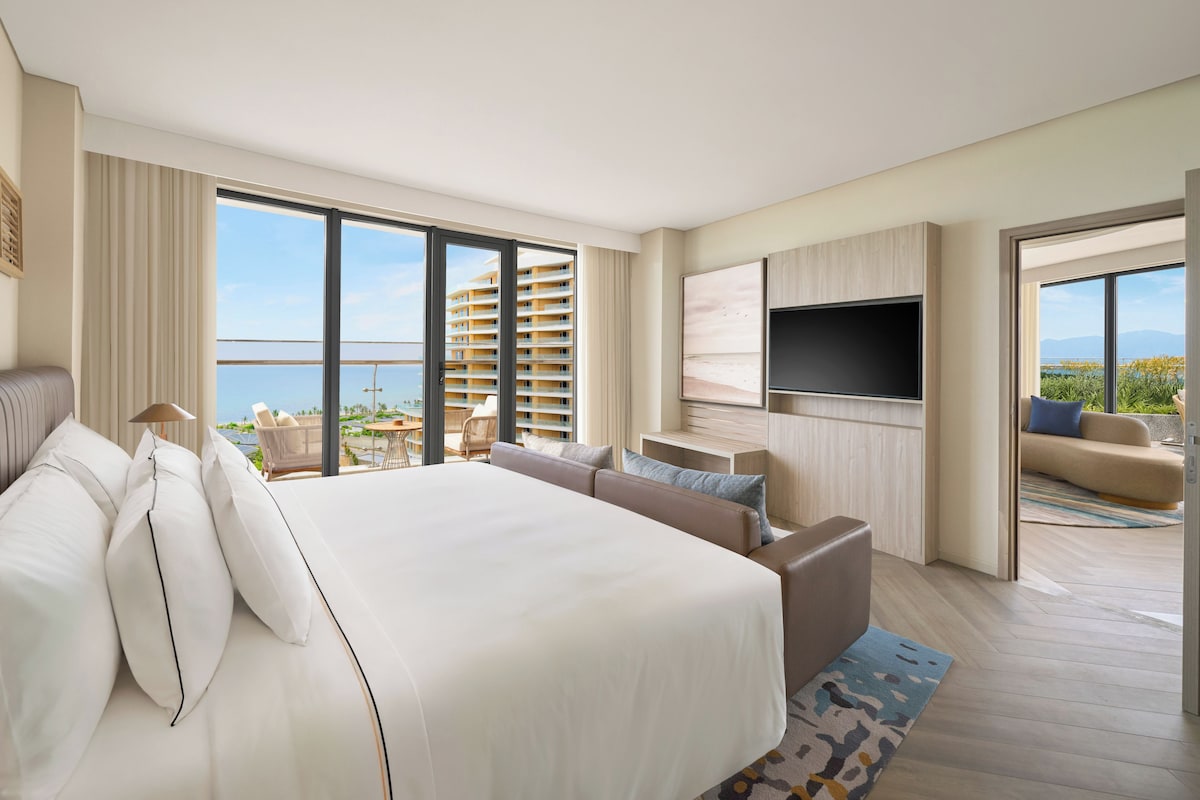 A hotel room with a large bed, a TV, a sofa, and a balcony with a view of the sea.