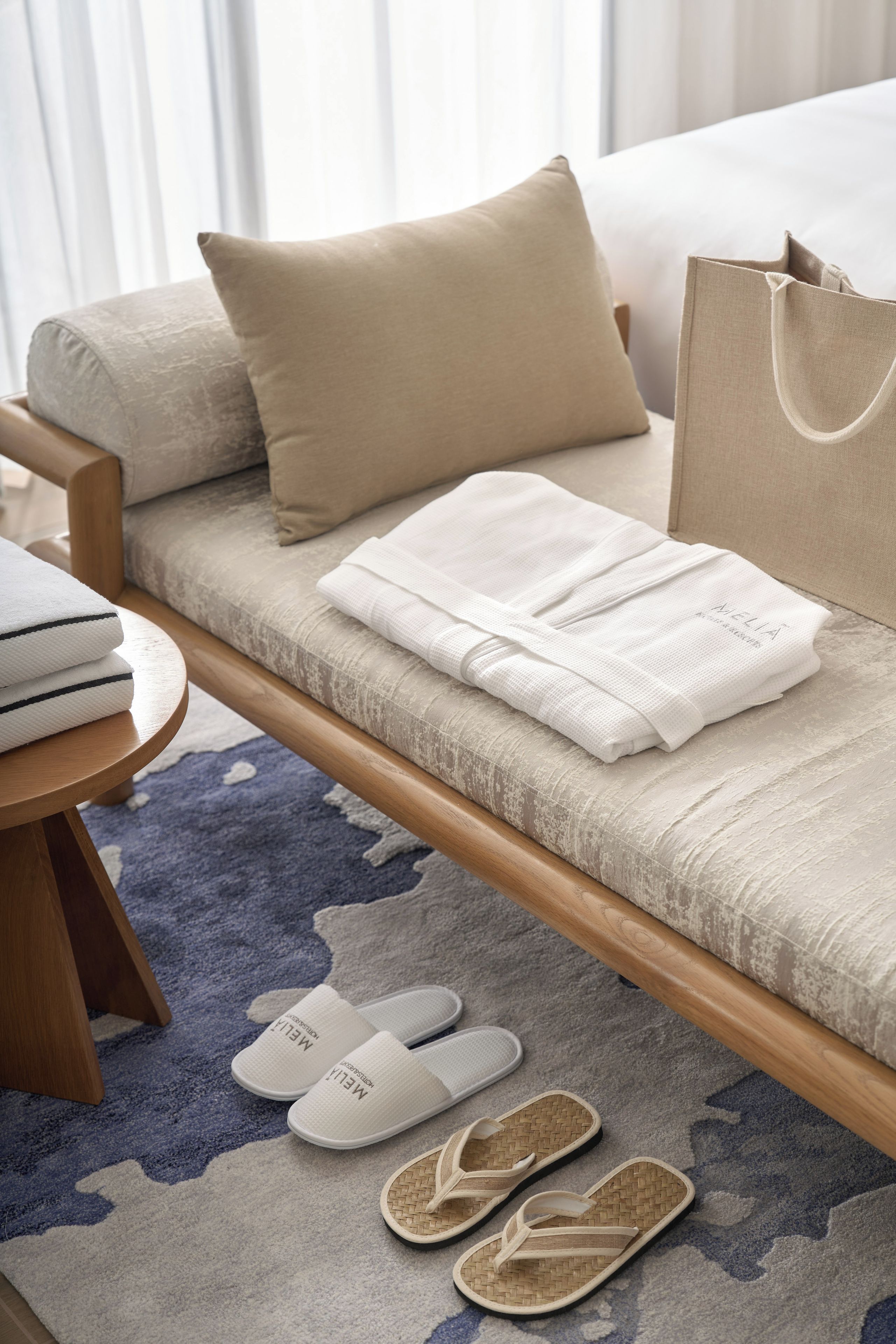 Una cama ordenada con almohadas, una toalla doblada, pantuflas y un bolso sobre una alfombra azul estampada.