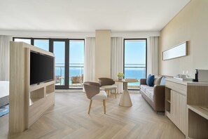 A modern hotel room with a flat-screen TV, a sofa, a dining table, and a balcony with a view of the sea. - Melia Serenity Cam Ranh Beach Resort (Cam Lam)
