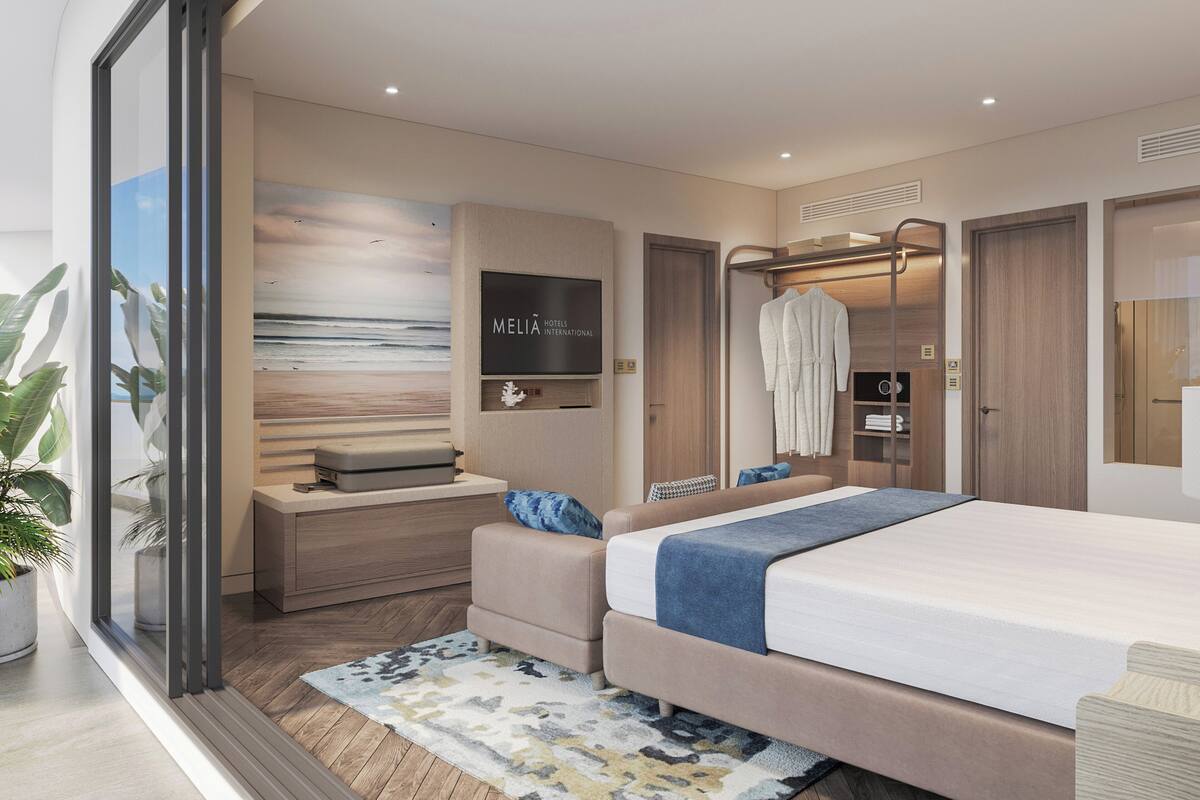 Ein modernes Hotelzimmer mit einem großen Bett, einer Sitzecke, einem Schreibtisch mit Koffer sowie einem an der Wand befestigten Fernseher und einem Wandbild mit Strandmotiv.