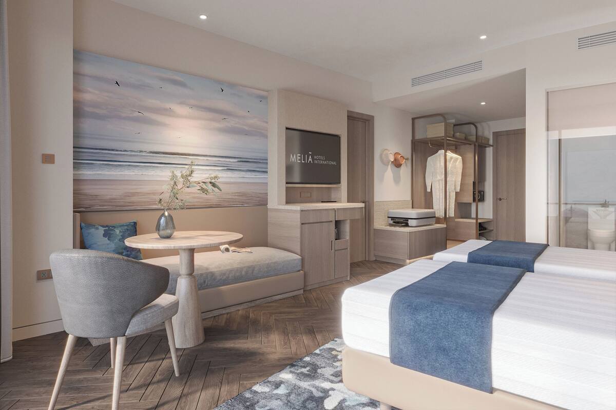 Ein modernes Hotelzimmer mit einem großen Gemälde einer Strandansicht, einem runden Tisch mit einer Vase, einer Couch, einem Bett mit blauer Bettwäsche und einem Kleiderschrank.