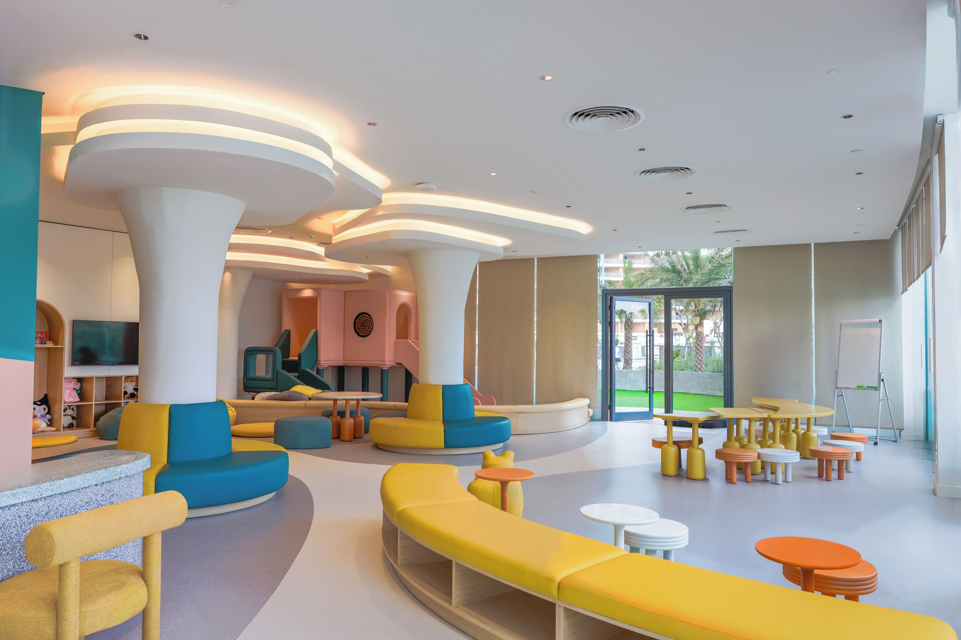 A brightly lit indoor space with modern furniture, including a yellow sofa, a pink play area, and multiple tables and chairs.