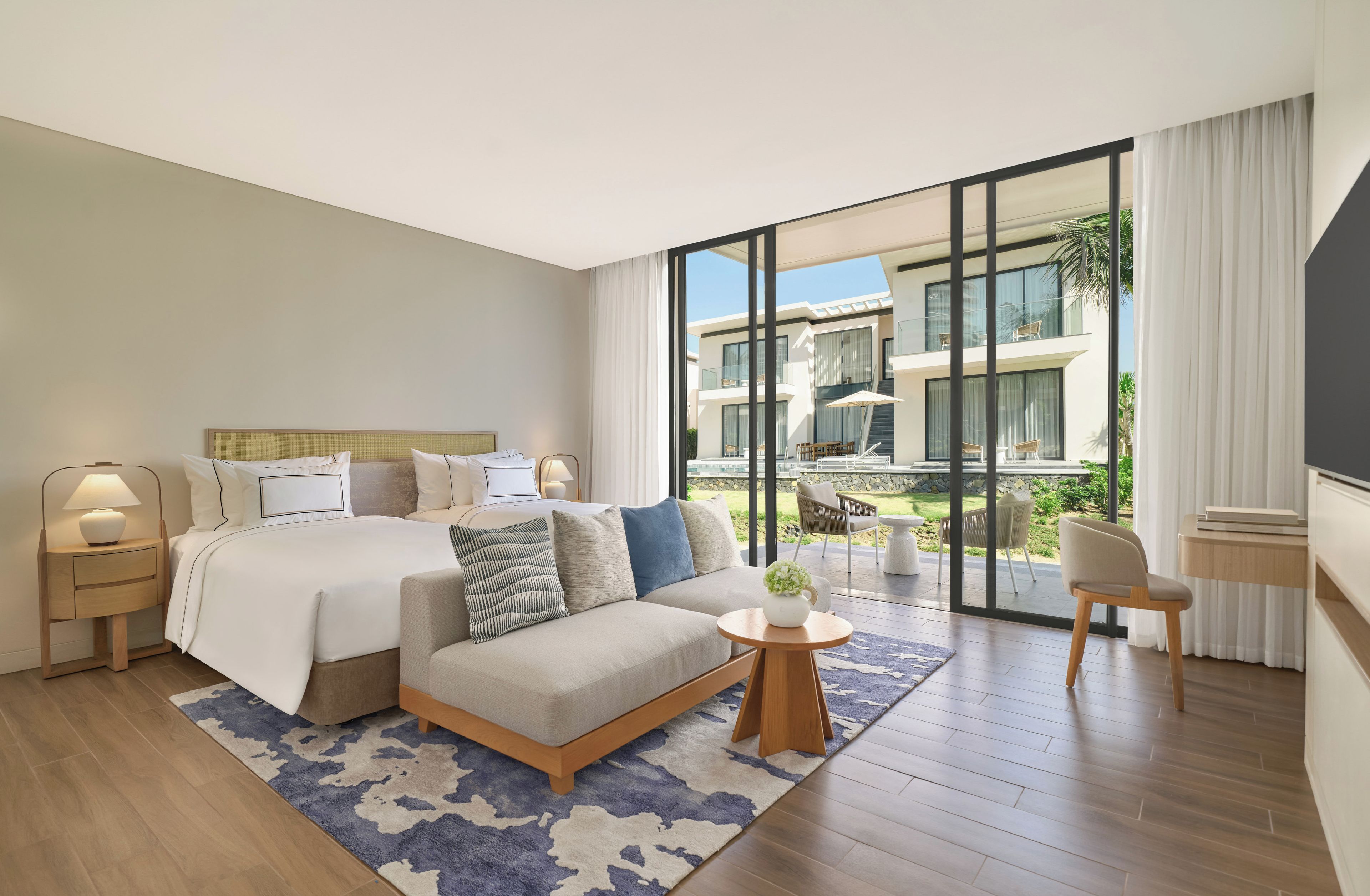 A modern hotel room with a large bed, a sofa, a small table, and a view of the outdoor balcony.