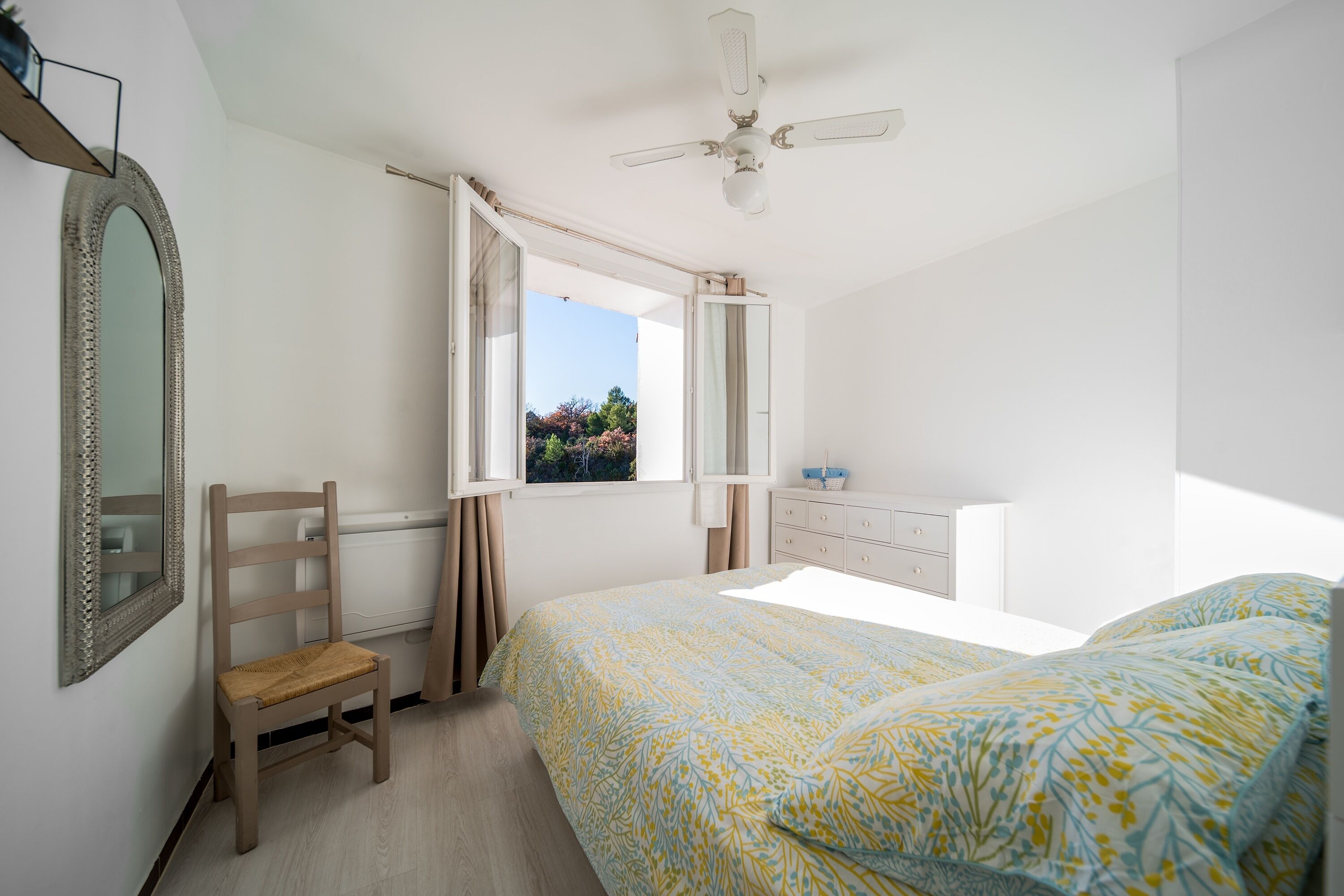 A bedroom with a bed, a chair, a dresser, a mirror, and a window with curtains.