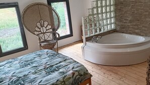 A bedroom with a large bed, a wooden floor, a white bathtub, and a window with a view of greenery.