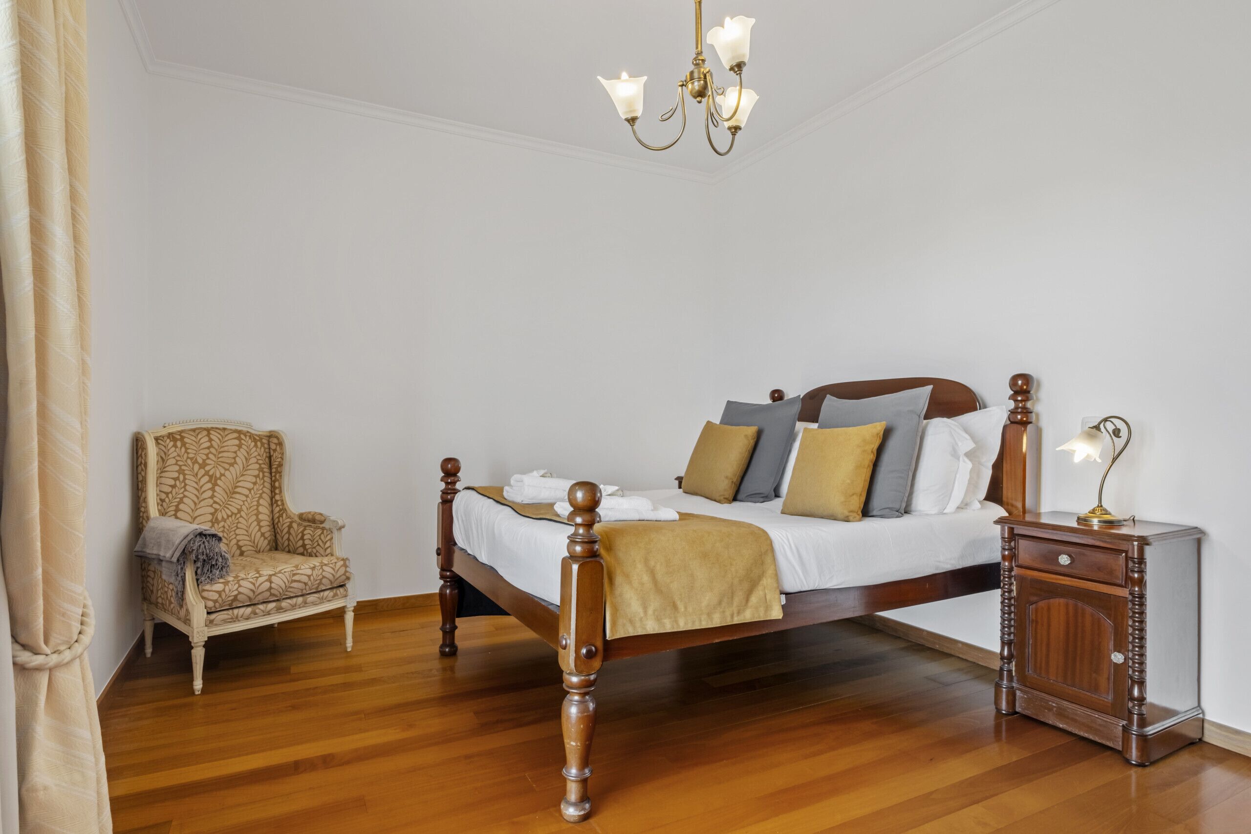A bedroom with a wooden bed, a nightstand with a lamp, a patterned armchair, and a chandelier.