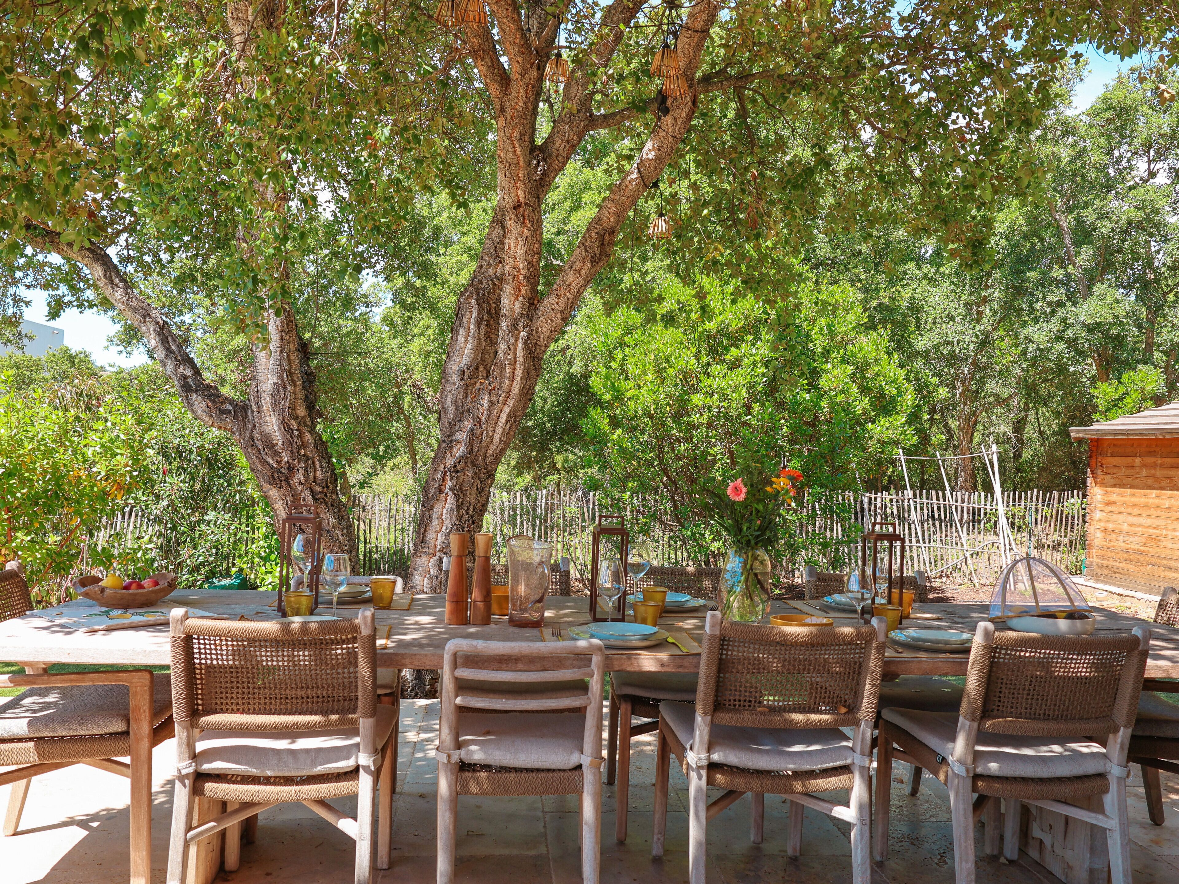 An outdoor dining area with wicker chairs and tables, surrounded by trees and a wooden fence.