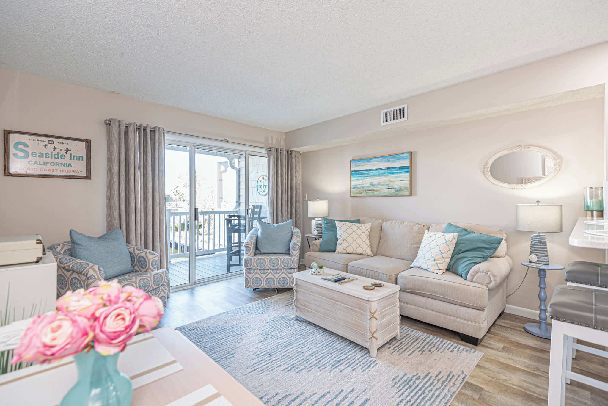 A living room with a sofa, armchairs, a coffee table, and a dining area. There is a large window with curtains, a painting on the wall, and a sign for Seaside Inn.