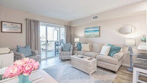 Una sala de estar con sofá, sillones, una mesa de centro y un comedor. Hay un ventanal con cortinas, un cuadro en la pared y un letrero que dice Seaside Inn.