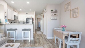 Una cocina moderna con armarios blancos, un microondas y un refrigerador. Hay una zona de comedor con una mesa y sillas de madera, y una isla de cocina con taburetes.