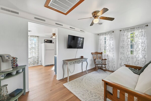A living room with a wooden floor, a ceiling fan, a TV, a wooden chair, a coffee machine, and a table.