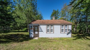 A small, single-story house with a brown roof and white siding, surrounded by trees and grass. - Cozy 1-bedroom cottage with waterfront views (Townsend)