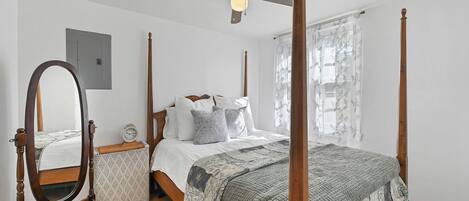 A four-poster bed with a wooden frame, a round mirror, a bedside table with a clock, and a window with sheer curtains.