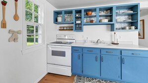 A kitchen with white and blue cabinets, a stove, and a window with a view of greenery. - Cozy 1-bedroom cottage with waterfront views (Townsend)