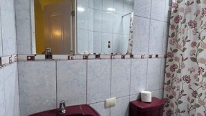 Une salle de bain avec un mur recouvert de carreaux de marbre, un lavabo rouge, un miroir et des toilettes agrémentées d’un rideau à motifs floraux.
