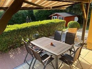 Eine Terrassesituation im Freien mit einem Holztisch und -stühlen, umgeben von einer Hecke und einem kleinen roten Gebäude im Hintergrund.