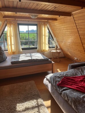 Ein Schlafzimmer mit schrägverdeckter Decke, einem Bett mit grau-weiß gestreifter Tagesdecke, einer roten Zudecke und Blick ins Grüne durch große Fenster.