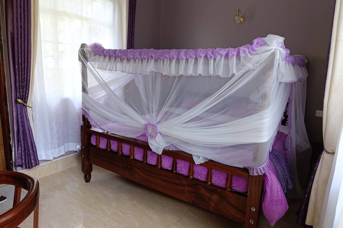 A wooden bed with a white and purple canopy, a wooden chair, and a window with purple curtains.