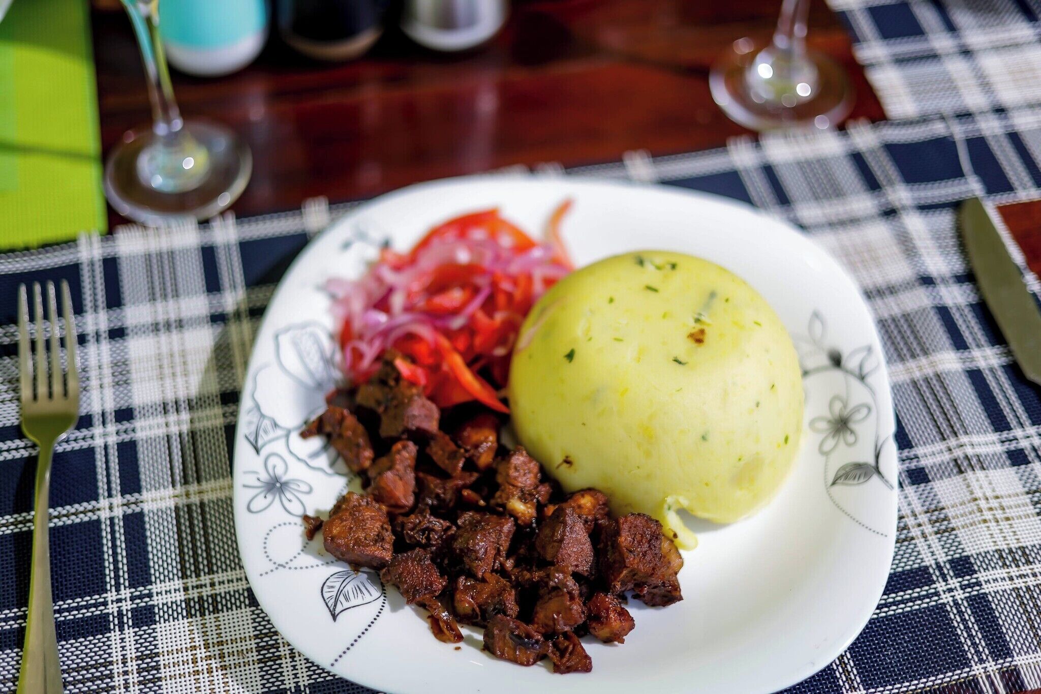 Ein Teller mit Fleisch, roten Zwiebeln und einem gekochten Kartoffel auf einer karierten Tischdecke.