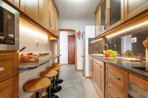 Eine moderne Küche mit Holzausstattung, einem Edelstahlkühlschrank und einer Arbeitsplatte aus Granit. Vier hölzerne Barhocker stehen an der Theke.