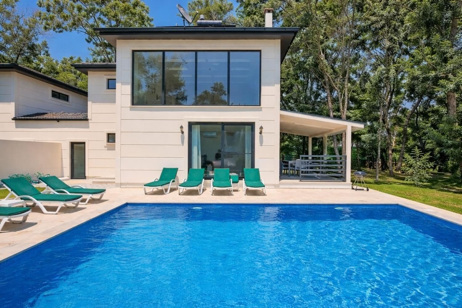 Een modern huis met twee verdiepingen, een zwembad, groene lounge fauteuils en een overdekt terras.