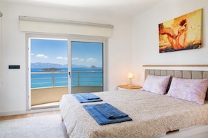 A bedroom with a bed, a nightstand, a lamp, a painting, and a balcony with a view of the sea and mountains.