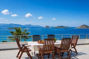 A terrace with a table set for four, overlooking a coastal view with mountains in the distance.