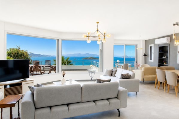 A spacious living area with a large sofa, a TV, and a balcony with a view of the sea.