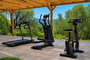 Três máquinas de exercício sob uma pérgola de madeira, com vista para árvores e colinas verdes.