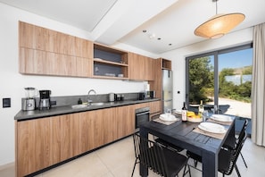 A modern kitchen with wooden cabinets, a dining table set for two, and a view of the outdoors. - Massari Hills Moon Villa (Archangelos)