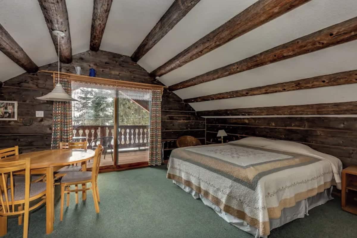 Um quarto de cabana acolhedor com teto de madeira, uma cama com manta listrada, uma mesa de jantar de madeira com cadeiras e vista para um deck exterior através de uma porta de vidro.