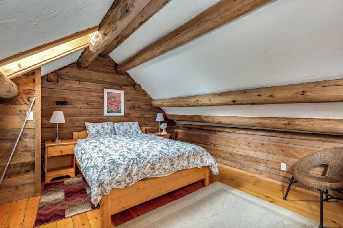 Um quarto de sótão acolhedor, com paredes de madeira, uma cama de madeira, uma mesa de cabeceira, uma cadeira e uma janela.