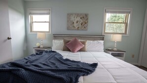 A bedroom with a bed, two bedside tables with lamps, a window with blinds, and a floral artwork on the wall.