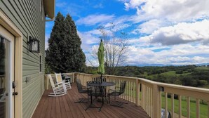 A wooden deck with a table and chairs, a green umbrella, and a view of a tree and landscape.