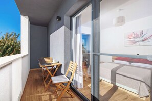 Ein Balkon mit einem Holztisch und Stühlen, ein Bett mit weißem Kopfteil und ein großes Fenster mit Blick auf Himmel und Bäume.