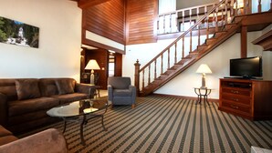 Un vaste salon comprenant un grand canapé, un fauteuil plus petit, une table basse en verre et un téléviseur posé sur un meuble en bois. Un escalier avec des rampes en bois mène à l’étage supérieur.