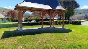 Ein hölzernes Pavillon mit kegelförmigem Dach, umgeben von einer gepflegten Rasenfläche und wolkenlosem Himmel.