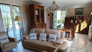 Ein Wohnzimmer mit einer Couch, einem Sessel, einem Holzschrank, einem Esstisch, einer Deckenleuchte und großen Fenstern.