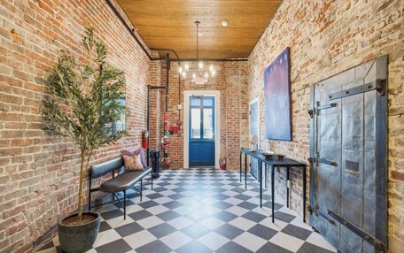 A hallway with a checkered floor, brick walls, a wooden ceiling, a bench, a potted plant, a table with a bowl, and a blue door at the end.