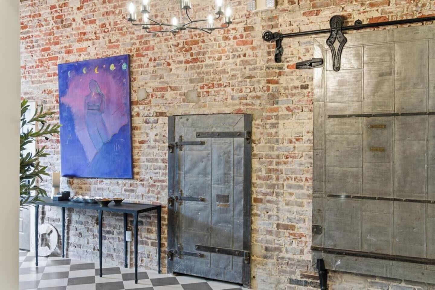 A room with exposed brick walls, a large abstract painting, a metal door, and a checkered floor.
