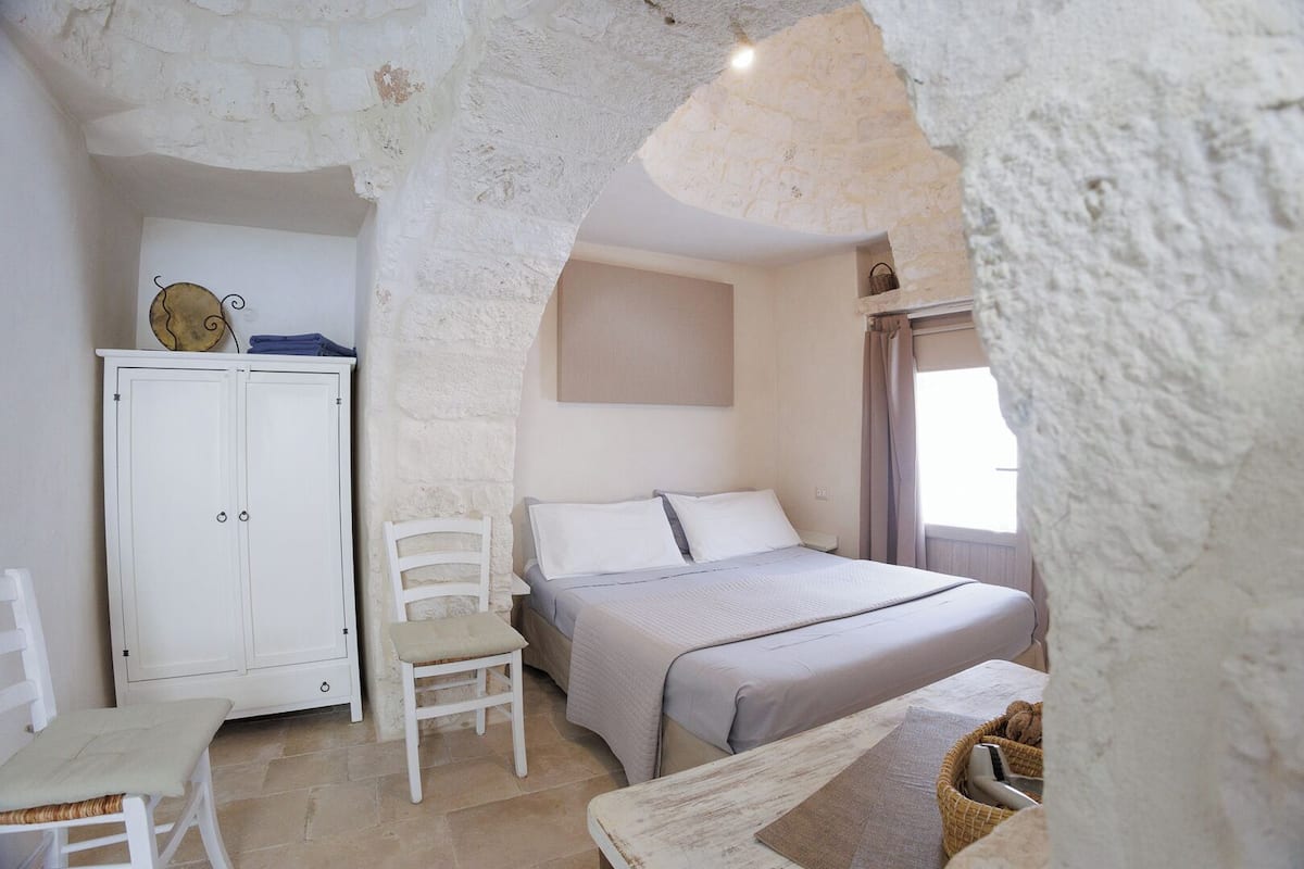 A bedroom with a stone wall, a bed, a chair, a desk, and a wardrobe.