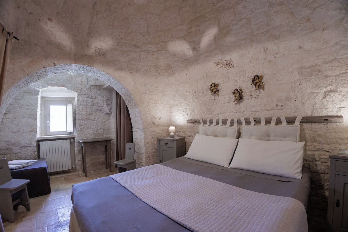 A bedroom with a stone wall, a bed with white linens, a small desk, and a window with curtains.
