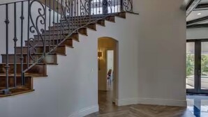 Ein großzügiger Innenraum mit einem Holzboden im Fischgrätmuster, einer Treppe mit aufwendiger Metallgeländerung und Blick auf eine Wohngegend durch große Fenster.