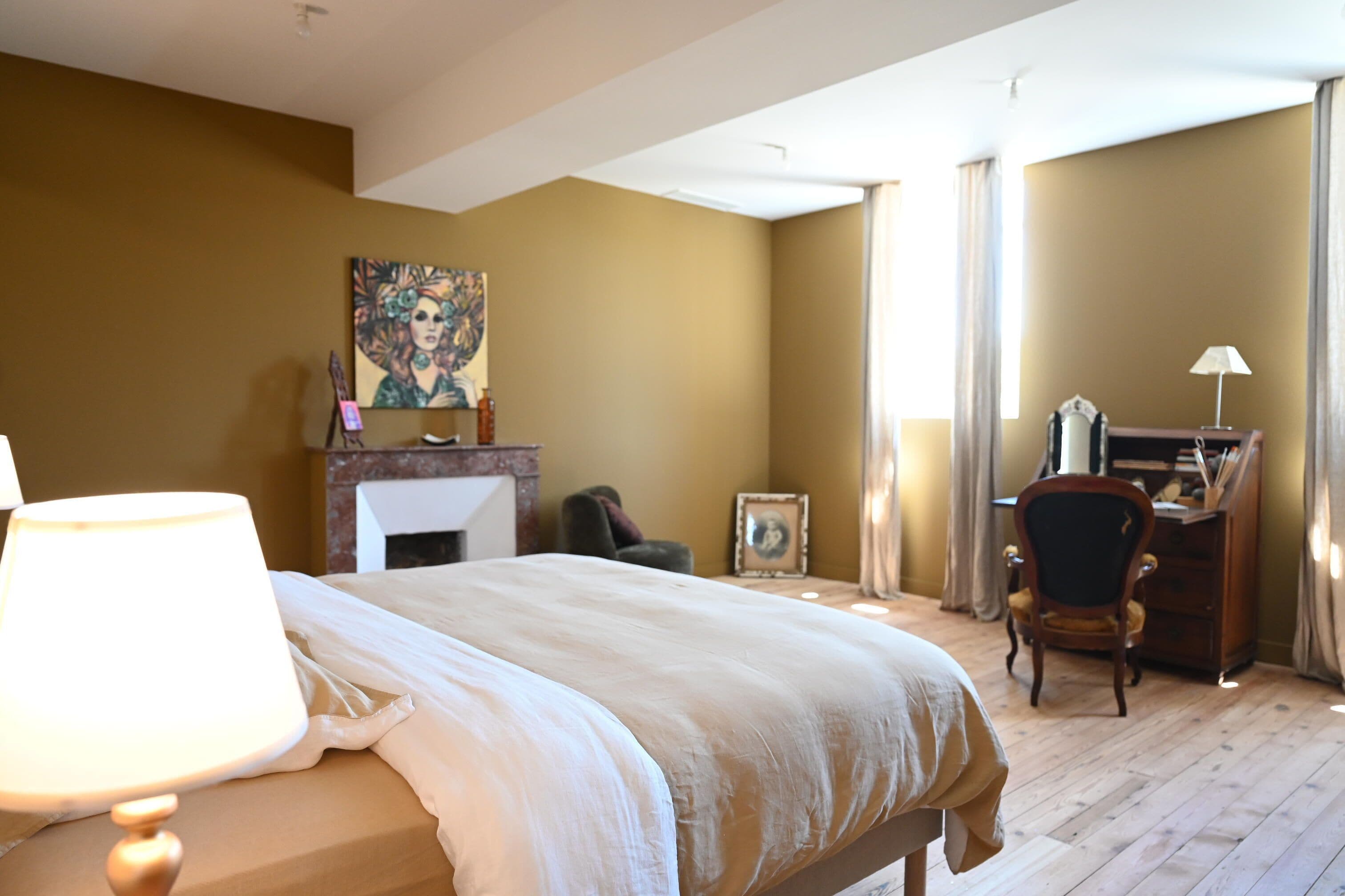 A hotel room with a large bed, a desk, a chair, a lamp, a fireplace, and a painting on the wall.